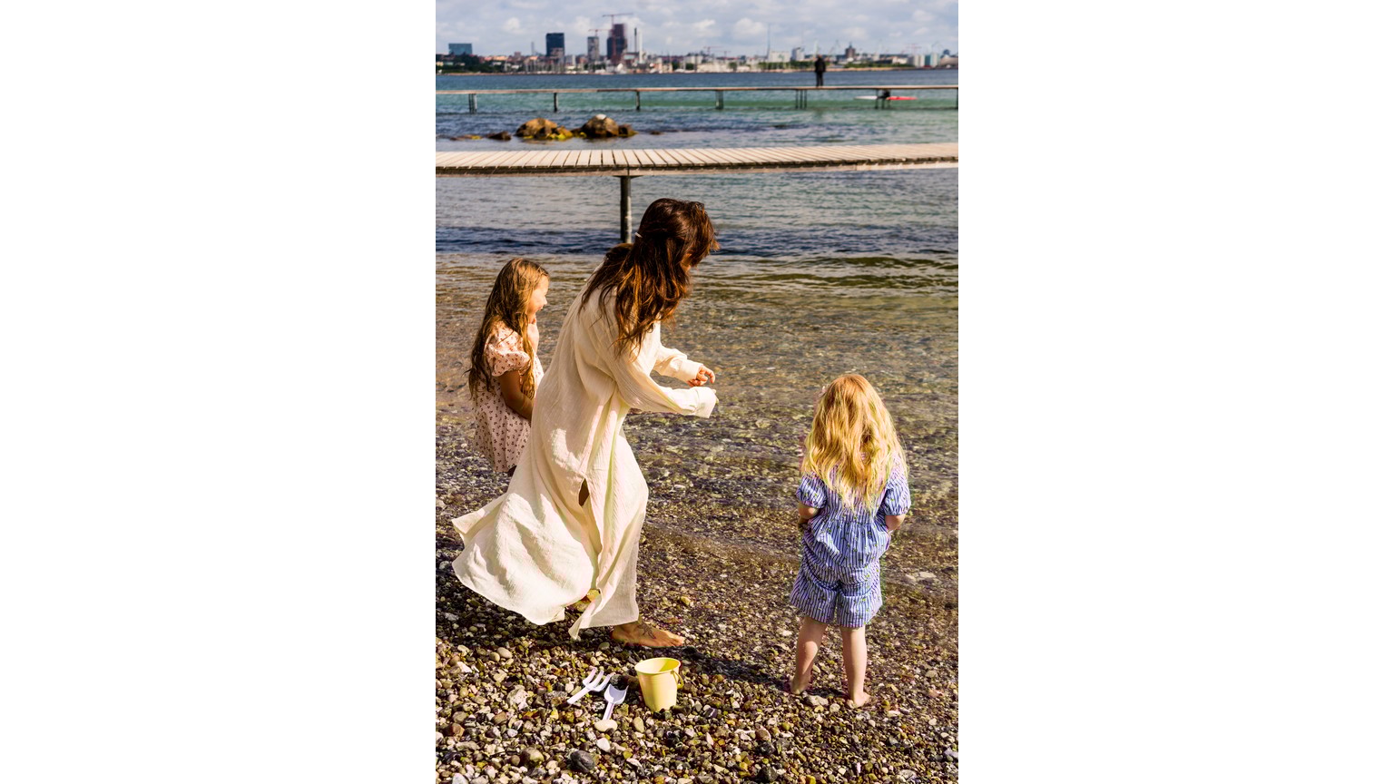 Ved vandkanten står en gruppe med ryggen til, mens de kaster sten i vandet med byens skyline i baggrunden.